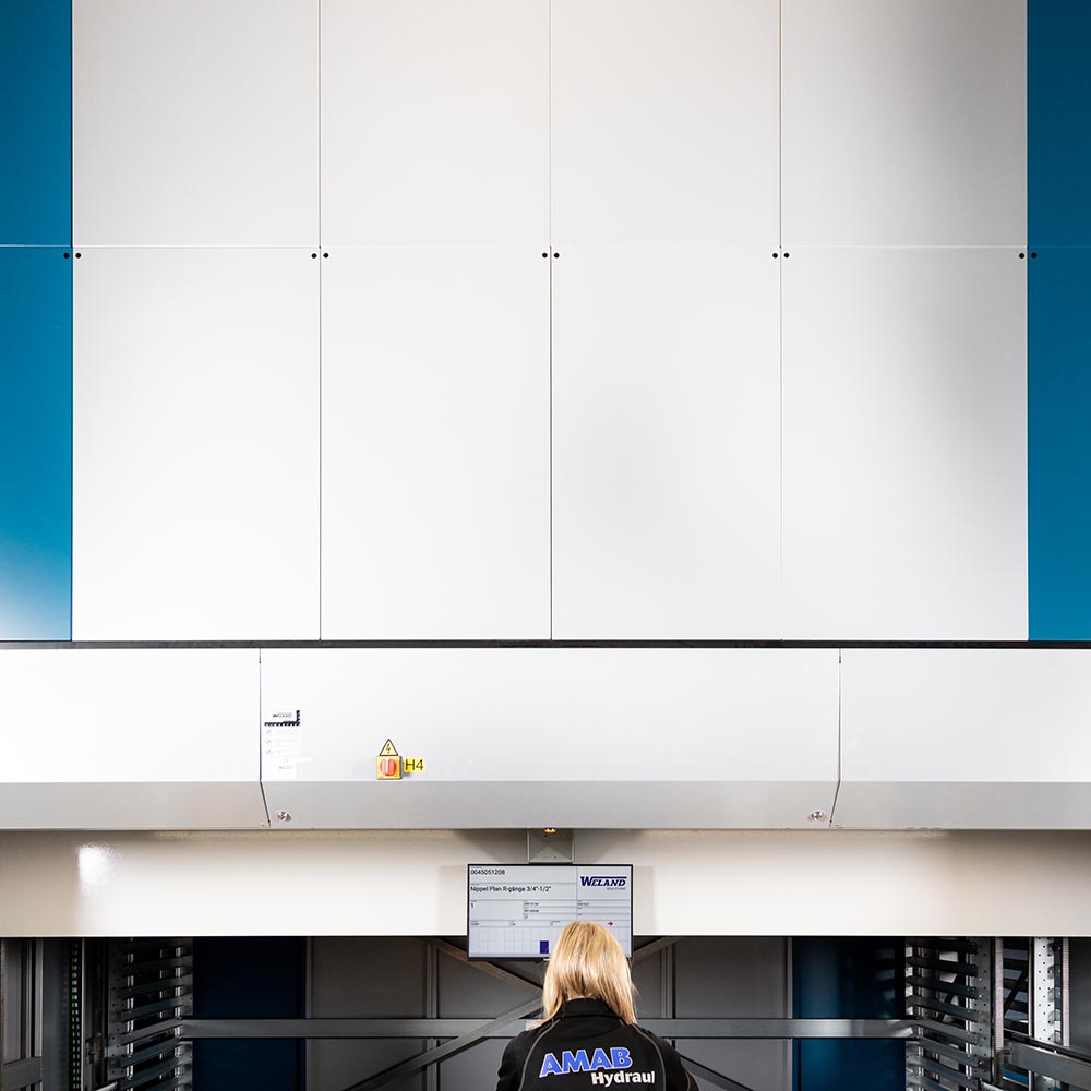 En kvinna sedd bakifrån står framför ett paternosterverk, eller även kallad en hissautomat. Hissautomaten är vit med blåa detaljer och står på ett lager.