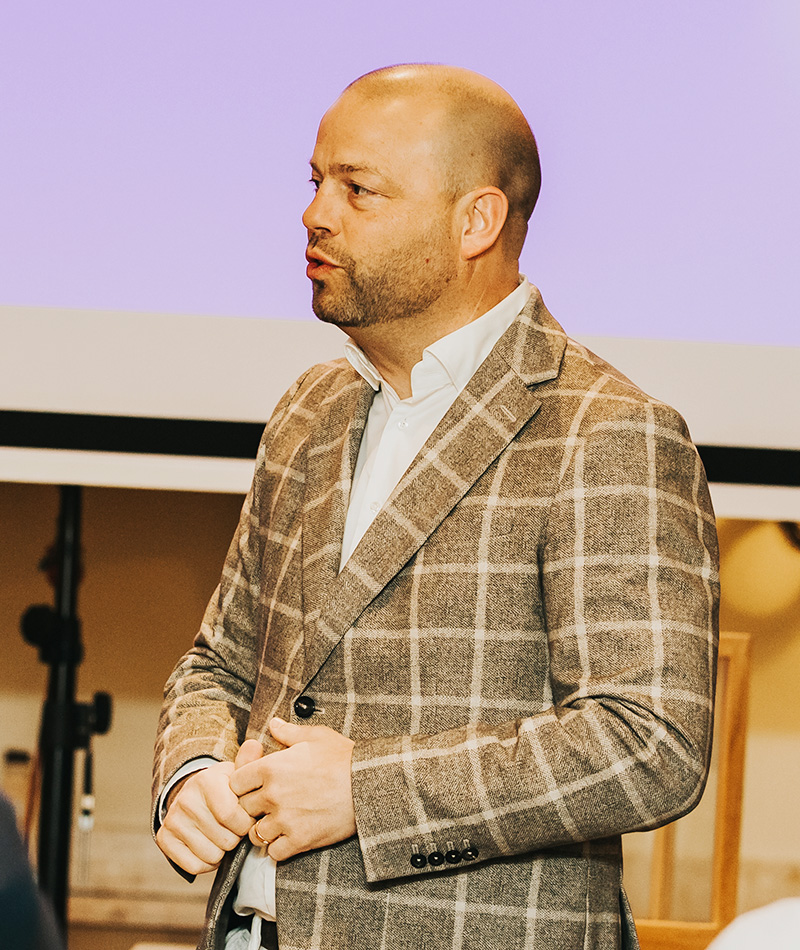 En man i rutig kavaj står framför en presentation med lila bakgrund, fångad i ett ögonblick av tal eller diskussion.