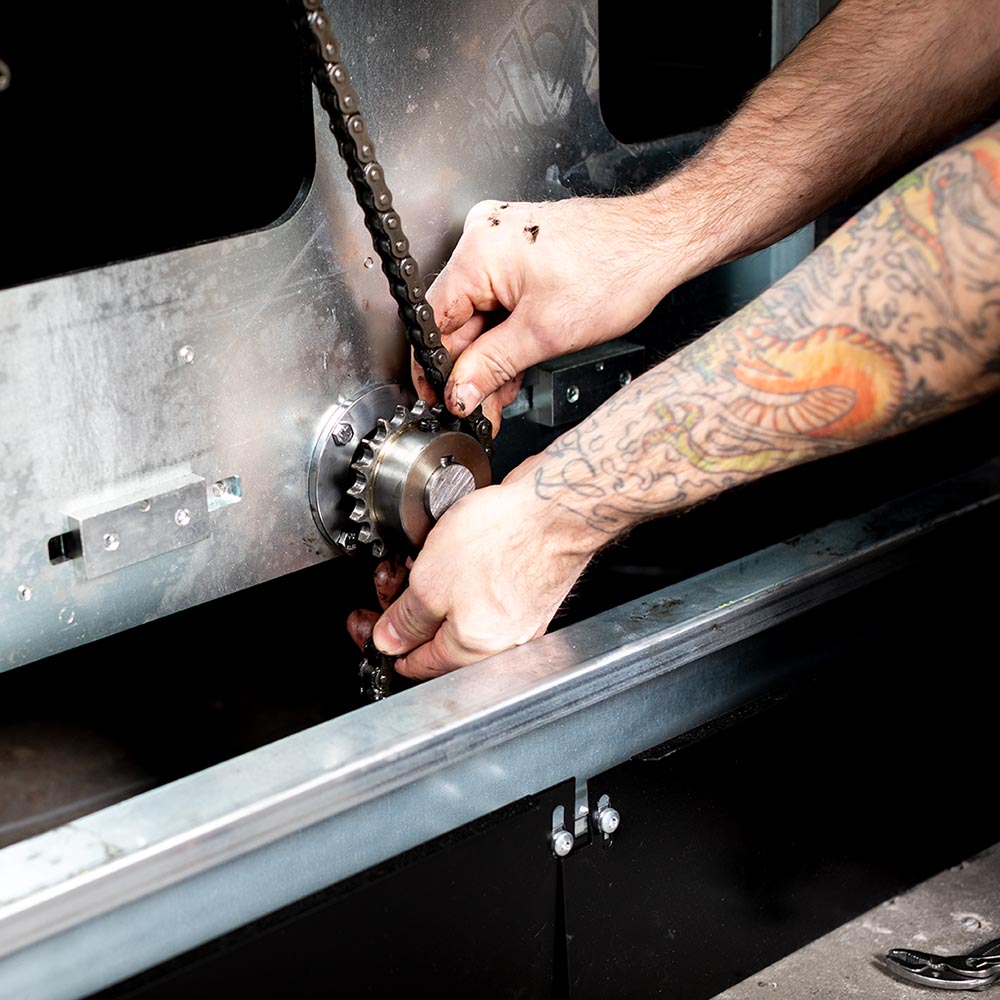 Två armar och händer som arbetar med underhåll av en hissautomat. Bilden visar hur man smörjer och sätter tillbaka kedjan på ett kugghjul på en hissautomat.