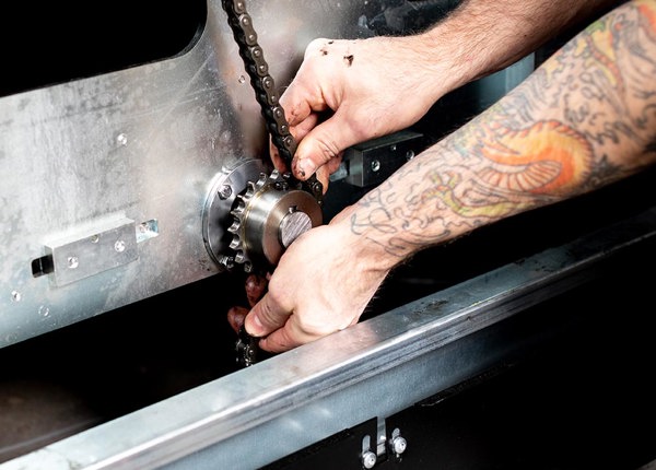 Två armar och händer som arbetar med underhåll av en hissautomat. Bilden visar hur man smörjer och sätter tillbaka kedjan på ett kugghjul på en hissautomat.