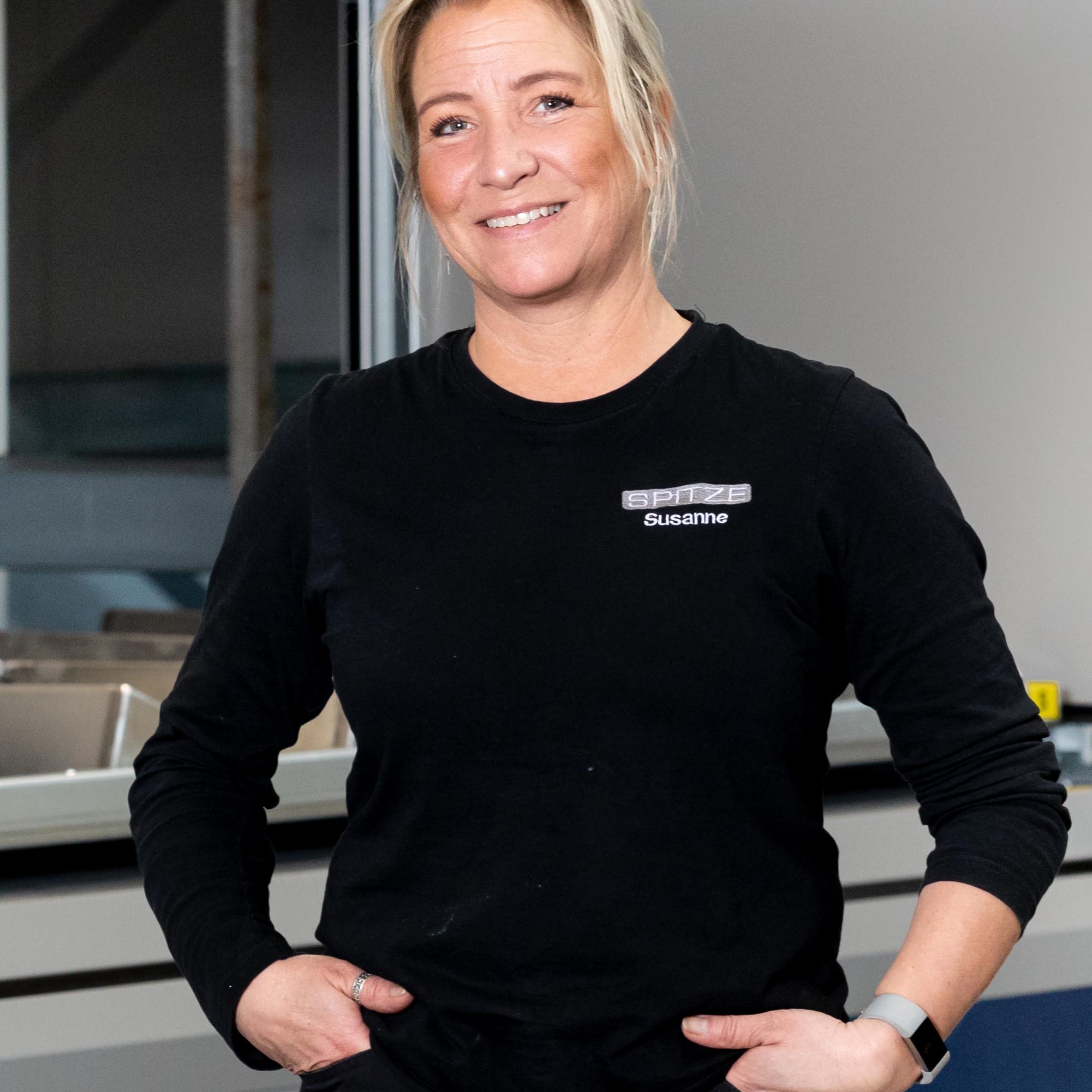 En kvinna som står vänd framåt. Hon är klädd i en svart tröja och står med händerna i fickorna och ser glad ut.