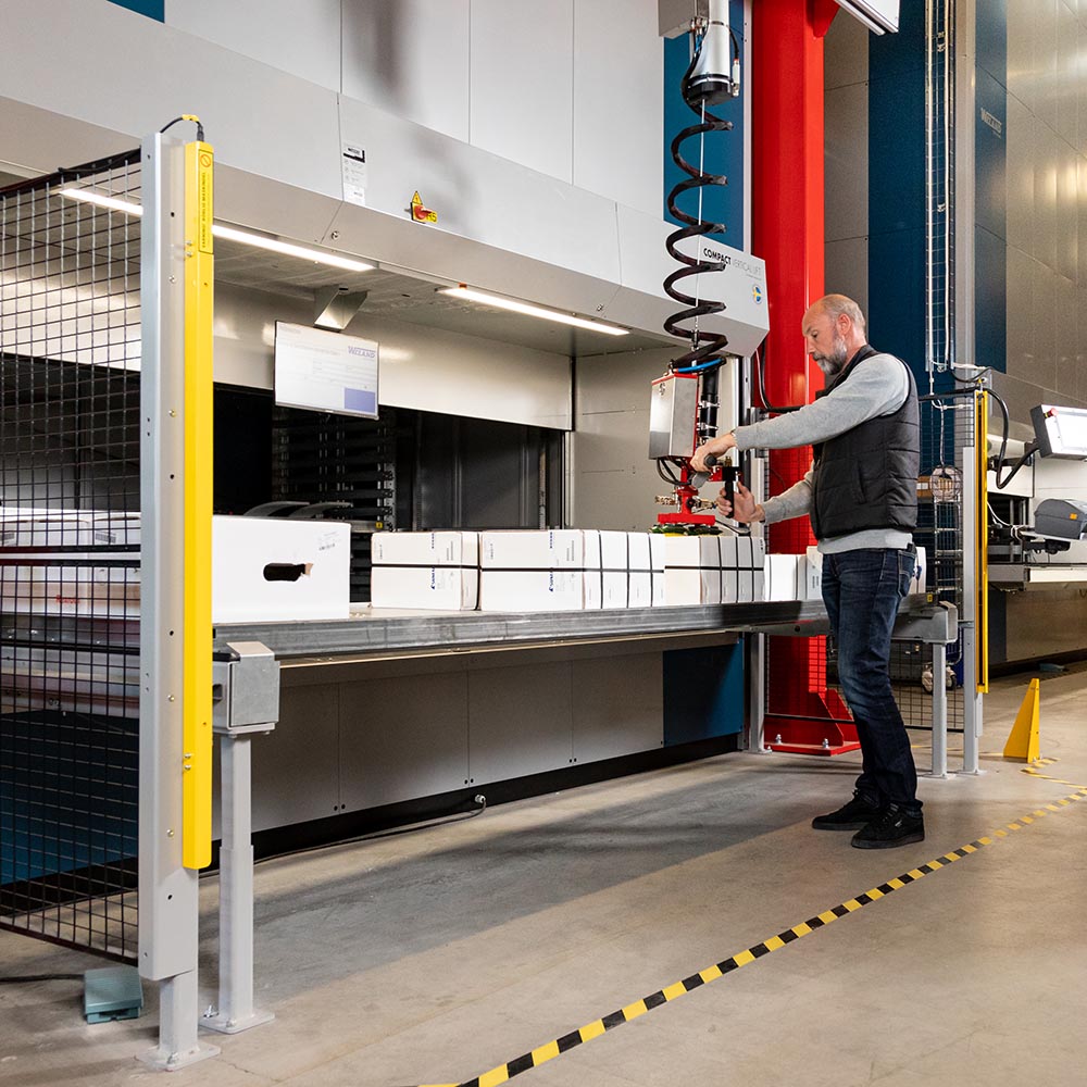 En man som står på ett lager och arbetar med en hissautomat av typen Compact. Detta händer på ett lager.