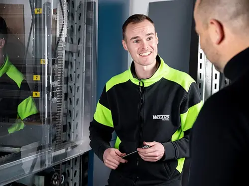 Två män samtalar vid en industriell maskin; den ena bär en svart och neongul arbetsjacka med Weland Solutions-logotypen och håller i ett verktyg.